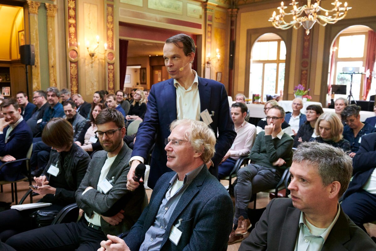 foto van groep mensen in een zaal en een man die de microfoon vasthoud tijdens een vraaggesprek